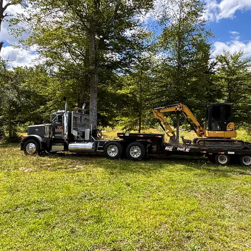 A tractor-trailer with a lowboy trailer carrying a compact excavator, parked in a mowed field under tall trees