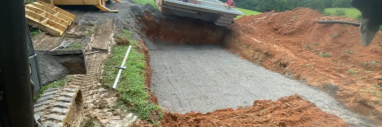 A fiberglass pool being lowered into a gravel-bedded excavation cut through red clay, with excavator tracks in the foreground
