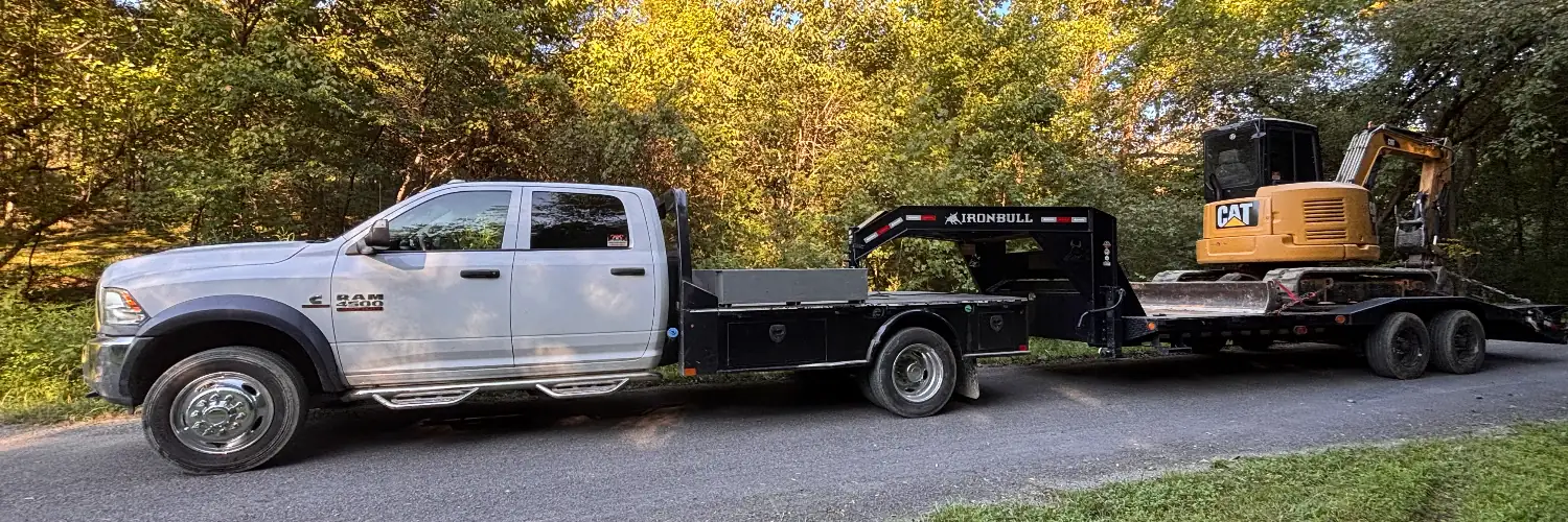 A white crew-cab flatbed pickup towing a gooseneck trailer loaded with a compact excavator on a tree-lined gravel road